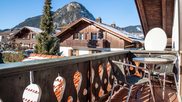 A wooden balcony with wicker chairs, a table, and a view of mountain houses.