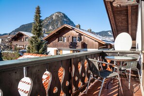 Un balcón de madera con sillas de mimbre, una mesa y vistas a casas de montaña.