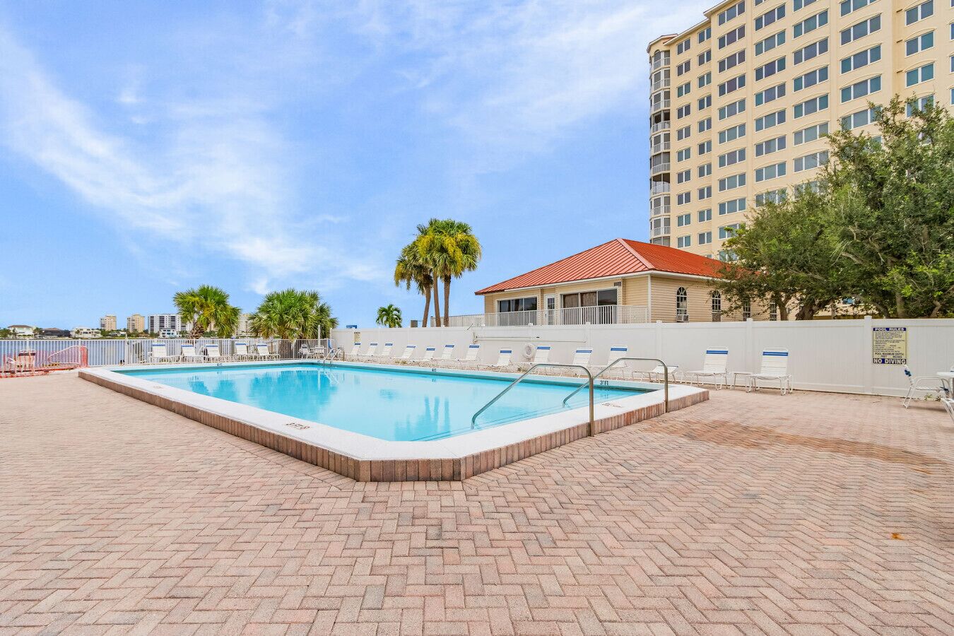 A rectangular swimming pool with a tiled deck, surrounded by lounge chairs and palm trees.