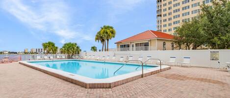 A rectangular swimming pool with a tiled deck, surrounded by lounge chairs and palm trees.