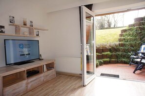 A modern living room with a television displaying 'Mami Kreisel' on the screen, wooden shelves with decorative items, a sliding glass door leading to an outdoor patio area with a table and chairs. - Peace and sea vacation with the Wyden family in Utersum on Föhr (Utersum)