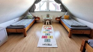 Um quarto pequeno e acolhedor com duas camas de madeira, uma janela com cortinas, uma mesinha e um tapete de jogos com cores do arco-íris.