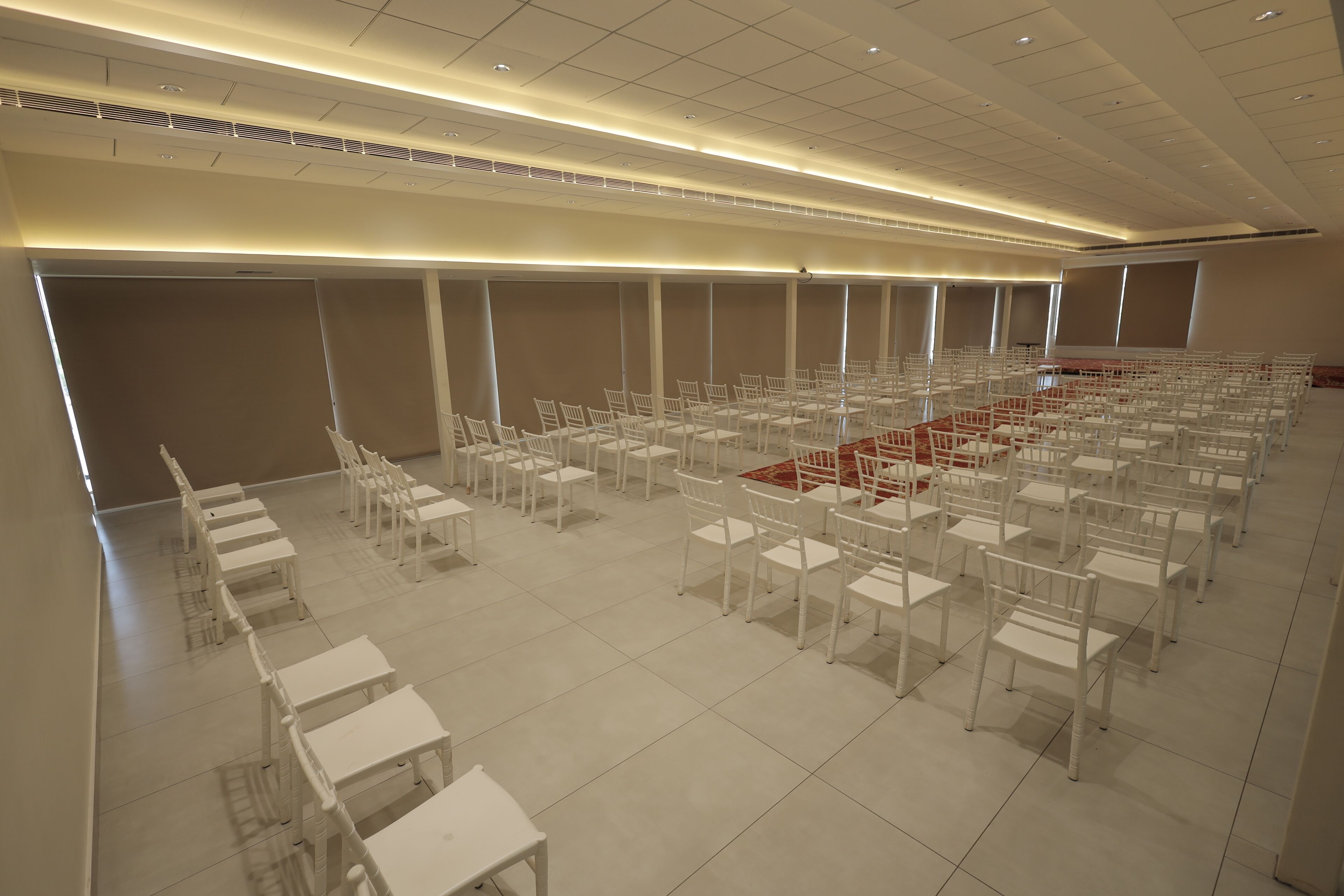 A large, empty conference room with rows of white chairs and tables, a red carpet, and a high ceiling with recessed lighting.