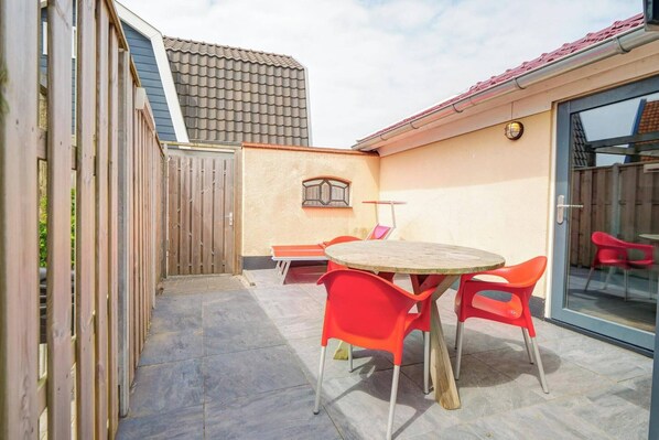 Un espace patio avec une table ronde en bois et des chaises en plastique rouge, une clôture en bois et un sol carrelé.