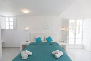 A hotel room with a bed, two bedside tables, a bench, a window, and a door. - Blue Island Hotel (Santorini)