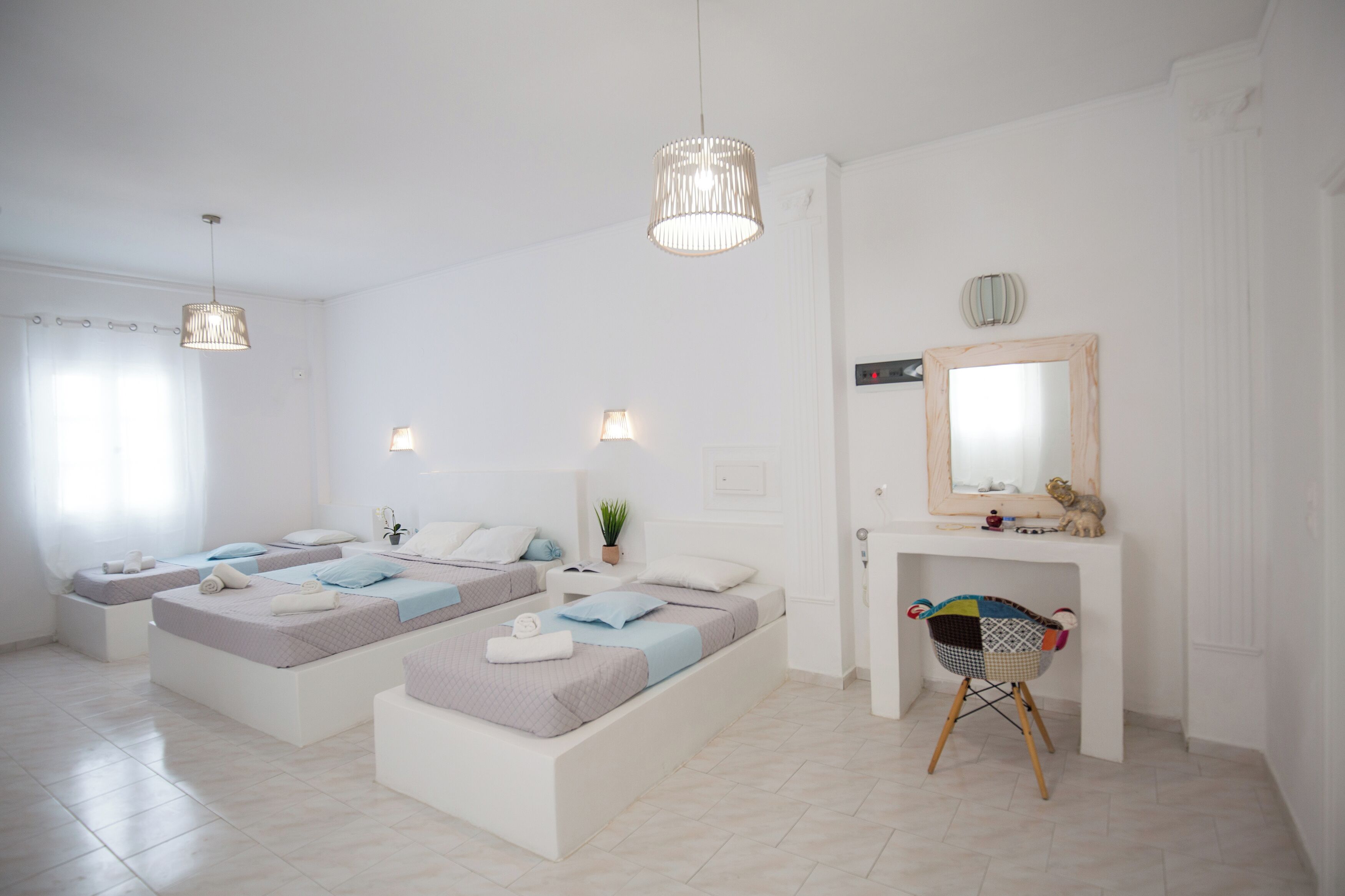 A modern hotel room with two single beds, a small desk, a chair, a mirror, and a hanging light fixture.