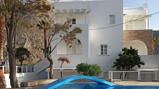 A clear swimming pool with a blue arch bridge, surrounded by white buildings and trees. - Blue Island Hotel (Santorini)