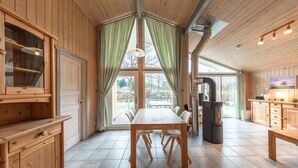 Un intérieur en bois comprenant une table à manger, des chaises, un poêle et une grande fenêtre avec des rideaux.