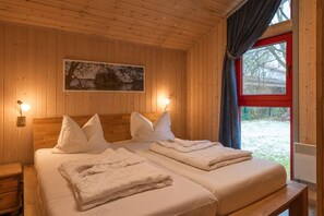 A wooden cabin room with two beds, a nightstand, a wall-mounted lamp, and a window with a view of a snowy landscape.