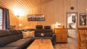 Interior acogedor de una cabaña con techo de madera, un sofá, una mesa de comedor y una cocina visible a través de una puerta abierta.