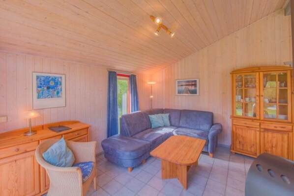 Ein gemütlicher Raum mit Holzboden, einem blauen Sofa, einem Holztisch, einem Korbstuhl und einer Kommode mit Glastüren.