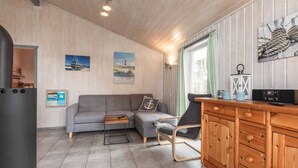 A cozy room with a grey sofa, a wooden cabinet, and a small table. There are two paintings on the wall, a lamp, and a window with curtains. - Ferienhaus mit Sauna im Ferienpark Mirow (Granzow)