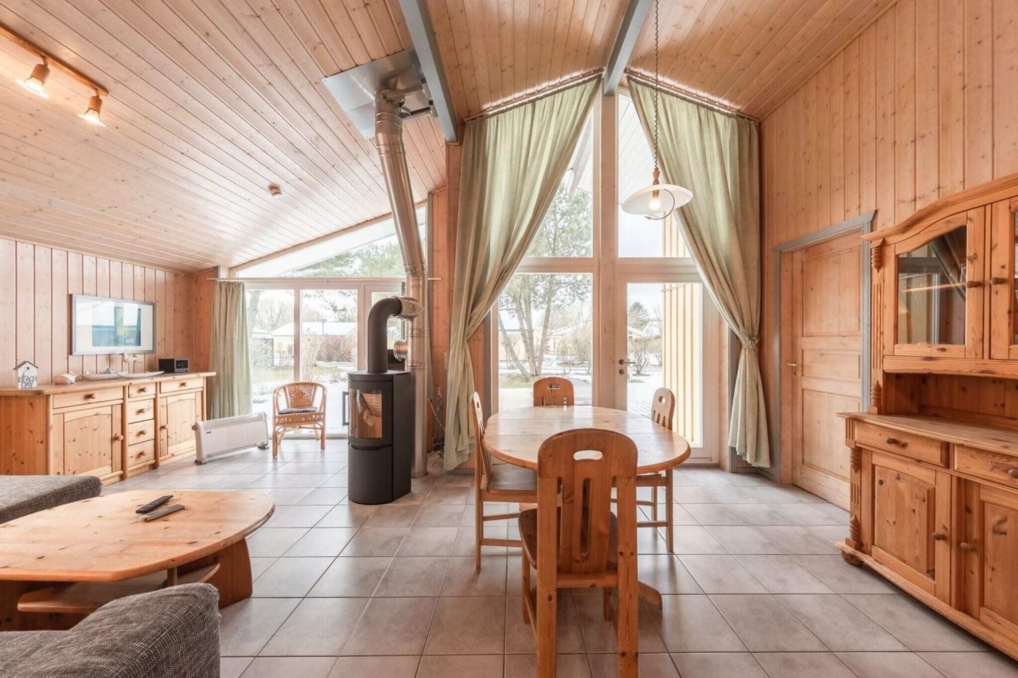 A wooden cabin interior with a dining table, chairs, a TV, a fireplace, and large windows.