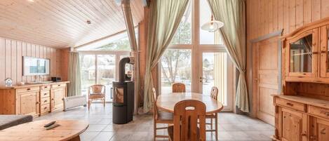 A wooden cabin interior with a dining table, chairs, a TV, a fireplace, and large windows.