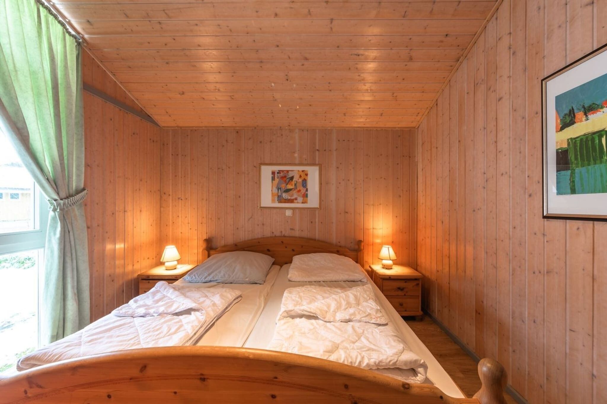 Een kamer met houten lambrisering, een bed, twee nachtkastjes met lampen, een lijst met een afbeelding aan de muur en een raam met gordijnen.