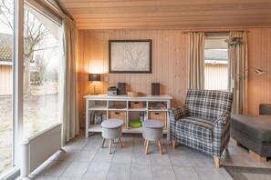 Une pièce chaleureuse avec un mur lambrissé en bois, un tableau encadré, une console blanche avec des haut-parleurs, un fauteuil à carreaux et un canapé gris.