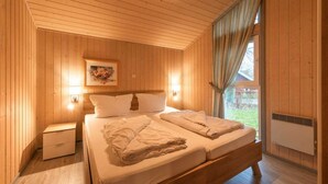 A wooden-paneled bedroom with a bed, bedside lamps, and a window with curtains. - Ferienhaus mit Sauna im Ferienpark Mirow (Granzow)