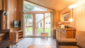 A cozy room with a wooden interior, a large sliding glass door leading to a garden, a sofa, a table, and a TV on a wooden stand. - Ferienhaus mit Sauna im Ferienpark Mirow (Granzow)