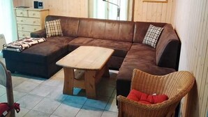 Un salon chaleureux avec un canapé d’angle marron, une table basse en bois, un fauteuil en osier et un petit meuble.