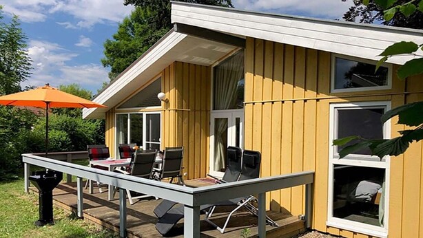Ein Holzhaus mit einer Terrasse, Sitzgelegenheiten im Freien und einem großen Fenster.