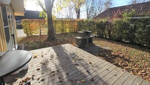 Eine Holzterrasse mit verstreuten Blättern, einer Steinsitzbank und einer Hecke in einem Garten.