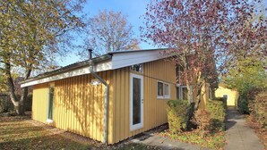 Ein kleines, eingeschossiges Holzhaus mit Haustür und Fenstern, umgeben von Bäumen und einem Bürgersteig.