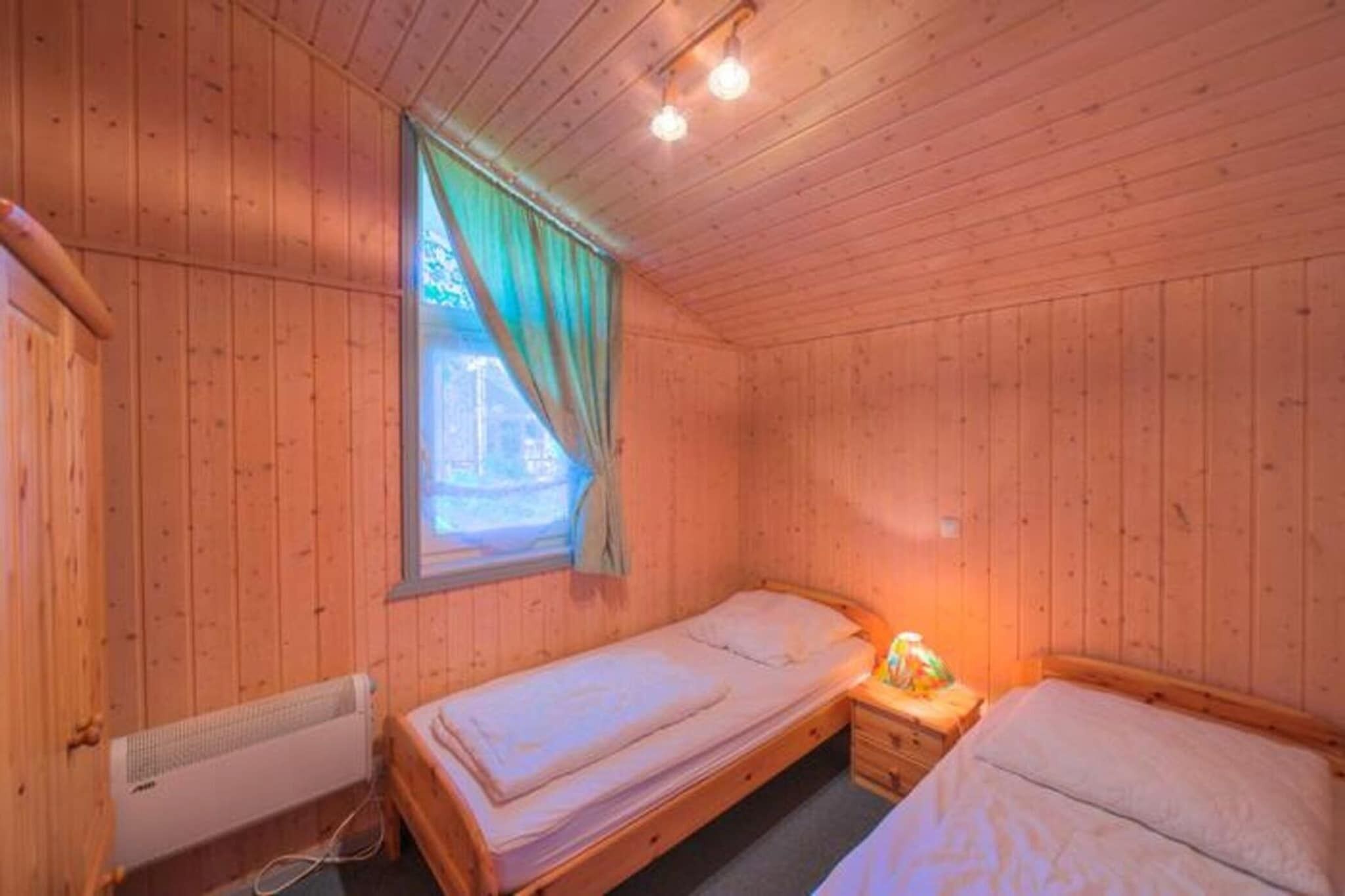 A wooden cabin room with two single beds, a window with curtains, a heater, and a nightstand with a lamp.