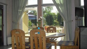 A dining area with wooden chairs and a table, large windows with curtains, and a television on the wall. - Ferienhaus mit Sauna im Ferienpark Mirow (Granzow)