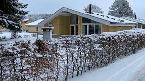 Ein modernes Haus mit Satteldach und großen Fenstern, umgeben von schneebedeckten Büschen und Bäumen.
