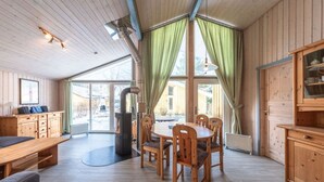 Ein Holzinterieur mit einem Esstisch und Stühlen, einem Kamin und großen Fenstern mit Vorhängen.
