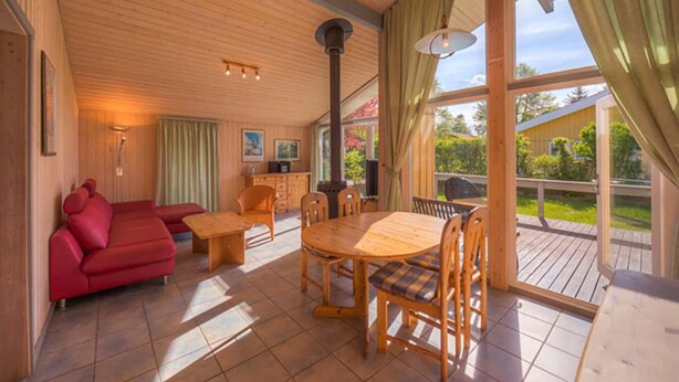 Ein gemütlicher Wohnbereich mit einem roten Sofa, einem Holzespeisetisch und Stühlen, großen Fenstern und Blick ins Freie.