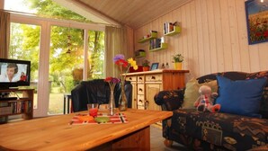 Ein gemütliches Wohnzimmer mit einem Holztisch, einer Couch und einem Fernseher. Es gibt Bücherregale und ein Fenster mit Blick auf Bäume.