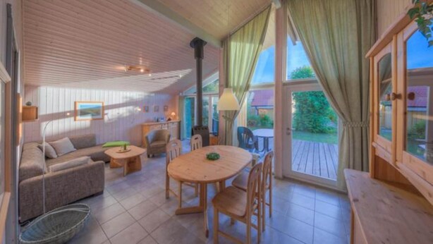Un salon chaleureux avec un canapé, une table ronde en bois et des chaises. Il y a une fenêtre avec des rideaux, une lampe et une vue sur la verdure à l’extérieur.