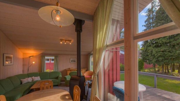 Ein gemütliches Blockhaus-Interieur mit Holzbalkendecke, einem grünen Sofa, einem Esstisch und Blick ins Freie durch große Fenster.