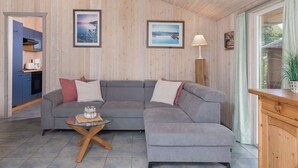 Ein modernes Wohnzimmer mit einem grauen Ecksofa, einem Glastisch und einem Holzschrank. An den Wänden hängen gerahmte Bilder und es gibt ein Fenster mit Vorhängen.