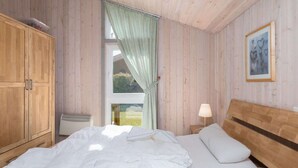 Ein Schlafzimmer mit Holzkopfteil, einem Bett mit weißer Bettwäsche, einem Nachttisch mit Lampe und einem Fenster mit Vorhängen.