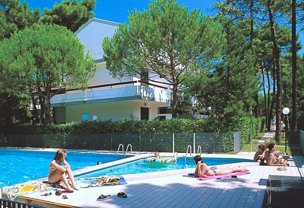 A swimming pool area with people relaxing and swimming, surrounded by trees and a building in the background.