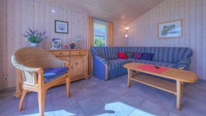 A cozy room with a wooden ceiling, a blue-striped sofa, a wicker chair, a wooden table, and a view through a window.