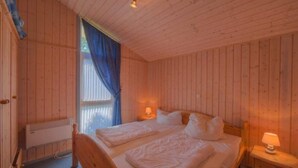 Camera da letto di una baita in legno con due letti, una finestra con tende blu e un comodino con una lampada.