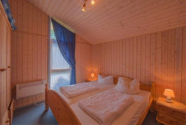A wooden cabin bedroom with two beds, a window with blue curtains, and a nightstand with a lamp.