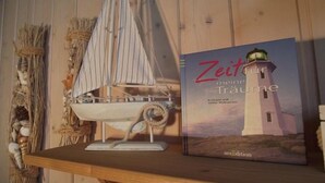 Ein Buch mit dem Titel "Zeit meiner Träume" mit einer Illustration eines Leuchtturms auf dem Cover, platziert auf einem Holzregal neben einem Modellsegelboot.