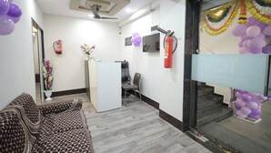 A reception area with a reception desk, a waiting area with a patterned sofa, a fire extinguisher, and a staircase with purple balloons. - The Grand Vinayak Hotel (Surat)