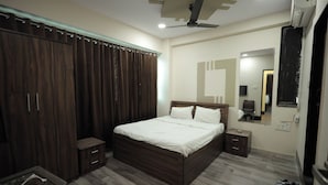 A bedroom with a wooden bed, a wardrobe, a nightstand, a mirror, and a ceiling fan. - The Grand Vinayak Hotel (Surat)