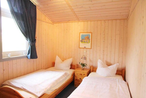 A room with two single beds, wooden walls, a window with curtains, a bedside table with a lamp, and a framed picture on the wall.