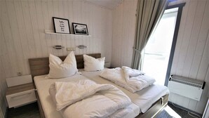 A neatly made double bed with white linens, a bedside table with a drawer, a wall-mounted shelf with two framed pictures, and a window with curtains.