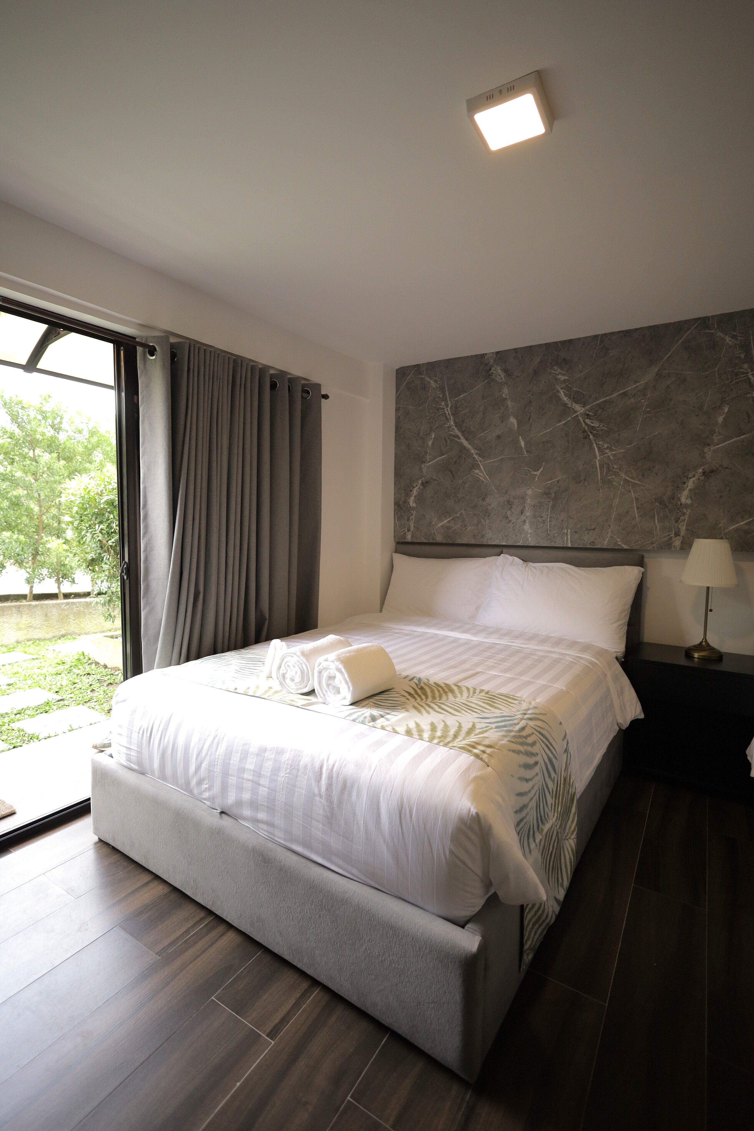 Un dormitorio moderno con una cama grande, cabecera de mármol, mesita de noche con lámpara y vista a zonas verdes.