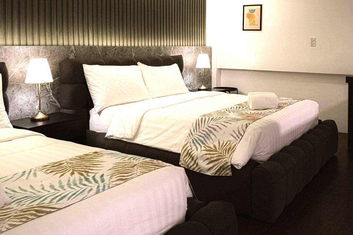 A hotel room with two beds, each with a headboard, bedside lamps, and a folded towel on the bed.