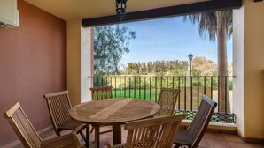 A covered patio with a round wooden table and four chairs, overlooking a fenced garden with trees and a clear sky.