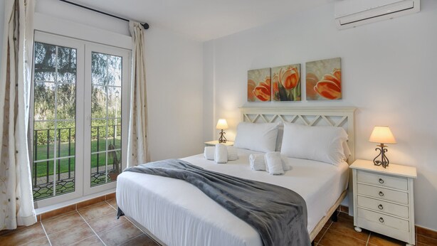 A hotel room with a bed, two bedside tables with lamps, a window with curtains, and two wall-mounted paintings.