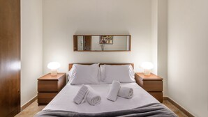 Un lit bien fait, avec du linge de lit blanc et deux serviettes enroulées, flanqué de deux tables de chevet en bois avec des lampes, dans une pièce dotée d’une porte en bois et d’un miroir au mur.
